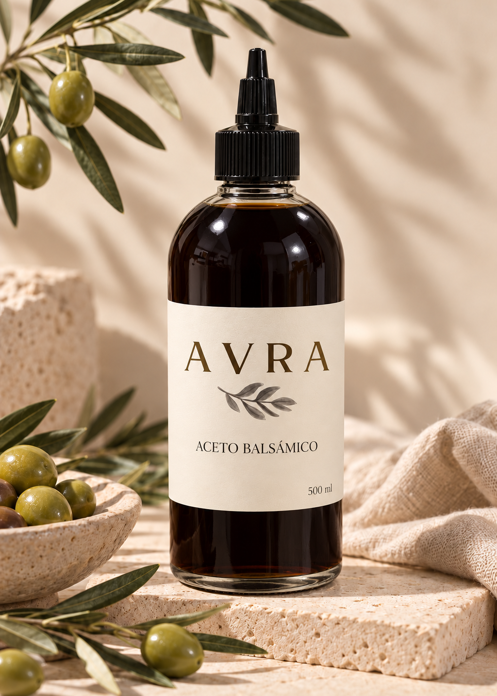 Botella de aceto balsámico AVRA de 500 ml con tapa dosificadora negra, junto a una rama de olivo y un bol de aceitunas